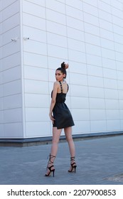 High End Fashion Image Of Tall Long Legs Slim Brunette Model Wearing Short Black Leather Dress, High Heels Summer Shoes. Bun Hairstyle. Outdoor Photo Shoot Over White Wall Background With Copy Space