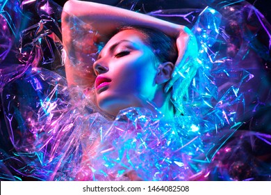 High Fashion model girl in colorful bright neon lights posing in studio through transparent film. Portrait of beautiful sexy woman in UV. Art design colorful make up. On colourful vivid background
