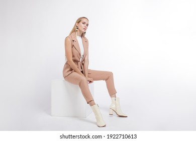 High fashion photo of a beautiful elegant young woman in a pretty beige suit, pants, vest, trousers, boots posing over white, soft gray background. Studio Shot, portrait. Model sits on a cube. Blonde - Shutterstock ID 1922710613