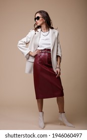 High Fashion Photo Of A Beautiful Elegant Young Woman In A Pretty White Leather Jacket, Red Long Skirt, Boots, Accessories, Stylish Sunglasses Posing Over Beige Background. Studio Shot. Portrait.