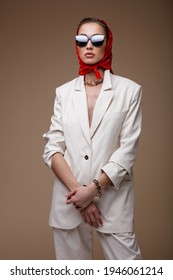 High Fashion Photo Of A Beautiful Elegant Young Woman In Pretty White Suit, Jacket, Trousers, Pants, Shoes, Red Scarf, Sunglasses, Accessories, Posing On Beige  Background. Studio Shot. Portrait.