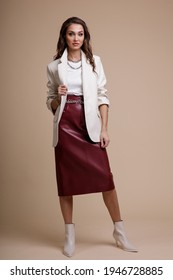 High Fashion Photo Of A Beautiful Elegant Young Woman In A Pretty White Leather Jacket, Red Long Skirt, Boots, Accessories Posing Over Beige Powdery Background. Studio Shot. Portrait.