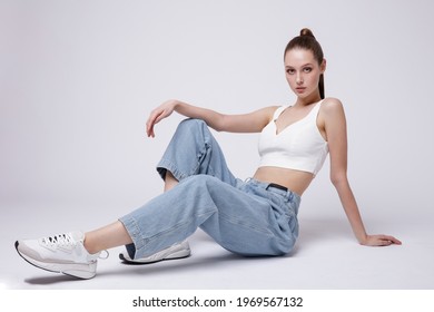 High fashion photo of a beautiful elegant young woman in a pretty white top and sneakers, blue denim jeans posing over white, soft gray background. Studio Shot. The model is sitting - Shutterstock ID 1969567132