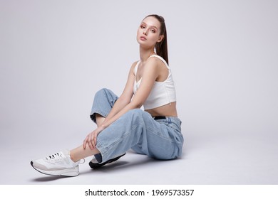 High fashion photo of a beautiful elegant young woman in a pretty white top and sneakers, blue denim jeans posing over white, soft gray background. Studio Shot. The model is sitting - Shutterstock ID 1969573357