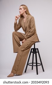 High Fashion Photo Of A Beautiful Elegant Young Woman In A Pretty Beige Suit, Button-down Pants, Jacket Posing Over White Background. Studio Shot. Slim Figure, Wide Clothes. Model Sitting On A Chair.