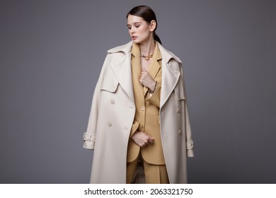 High fashion photo of a beautiful elegant young woman in a pretty white coat, beige sand suit, jacket, pants, trousers, accessories posing on gray background. Studio Shot. Slim figure. 