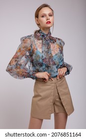 High fashion photo of a beautiful elegant young woman in a pretty  blue transparent blouse with a floral pattern, a bow on the neck, short beige shorts similar to a skirt posing over white background