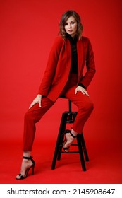 High Fashion Photo Of A Beautiful Elegant Young Woman In A Pretty Red Suit, Jacket, Pants, Trousers, Black Blouse Posing On Background. Monochrome, Total Red. Model Sits On Chair