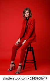 High Fashion Photo Of A Beautiful Elegant Young Woman In A Pretty Red Suit, Jacket, Pants, Trousers, Black Blouse Posing On Background. Monochrome, Total Red. Model Sits On Chair