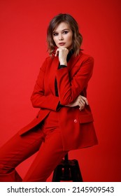 High Fashion Photo Of A Beautiful Elegant Young Woman In A Pretty Red Suit, Jacket, Pants, Trousers, Black Blouse Posing On Background. Monochrome, Total Red. Model Sits On Chair