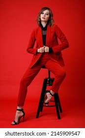 High Fashion Photo Of A Beautiful Elegant Young Woman In A Pretty Red Suit, Jacket, Pants, Trousers, Black Blouse Posing On Background. Monochrome, Total Red. Model Sits On Chair