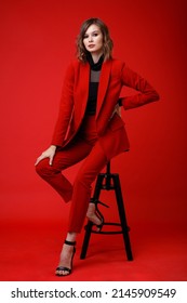 High Fashion Photo Of A Beautiful Elegant Young Woman In A Pretty Red Suit, Jacket, Pants, Trousers, Black Blouse Posing On Background. Monochrome, Total Red. Model Sits On Chair