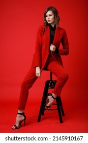 High Fashion Photo Of A Beautiful Elegant Young Woman In A Pretty Red Suit, Jacket, Pants, Trousers, Black Blouse Posing On Background. Monochrome, Total Red. Model Sits On Chair