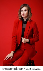 High Fashion Photo Of A Beautiful Elegant Young Woman In A Pretty Red Suit, Jacket, Pants, Trousers, Black Blouse Posing On Background. Monochrome, Total Red. Model Sits On Chair