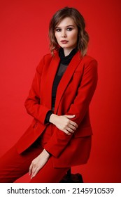 High Fashion Photo Of A Beautiful Elegant Young Woman In A Pretty Red Suit, Jacket, Pants, Trousers, Black Blouse Posing On Background. Monochrome, Total Red. Model Sits On Chair