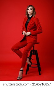 High Fashion Photo Of A Beautiful Elegant Young Woman In A Pretty Red Suit, Jacket, Pants, Trousers, Black Blouse Posing On Background. Monochrome, Total Red. Model Sits On Chair