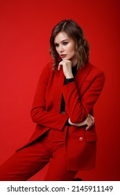 High Fashion Photo Of A Beautiful Elegant Young Woman In A Pretty Red Suit, Jacket, Pants, Trousers, Black Blouse Posing On Background. Monochrome, Total Red. Model Sits On Chair