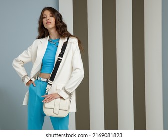 High Fashion Photo Of A Beautiful Elegant Young Asian Woman In A Pretty White Jacket, Blazer, Aqua Blue Top, Pants, Trousers, Accessories, Handbag. Striped Corrugated Wall. Studio Shot. 