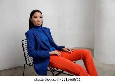 High fashion photo of a beautiful elegant young asian woman in pretty blue jacket, blazer, orange pants, trousers. White textured rounded wall. Model sits on chair