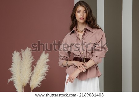 High fashion photo of a beautiful elegant young asian woman in pretty rosy brown shirt, belt, necklace, bracelets, white cream long skirt. Green striped Corrugated Wall, dry branches.