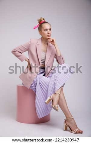 High fashion photo of a beautiful elegant young woman in pretty pink jacket, blazer, lilac lavender skirt posing on white, soft gray background. Slim Figure, Blonde. Model sits on cylinder, cube