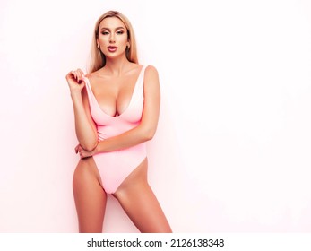 High fashion portrait of young beautiful sexy woman. Carefree model wearing pure pink lingerie with big breasts. Hot tanned blonde posing near wall in studio in summer swimwear bathing suit