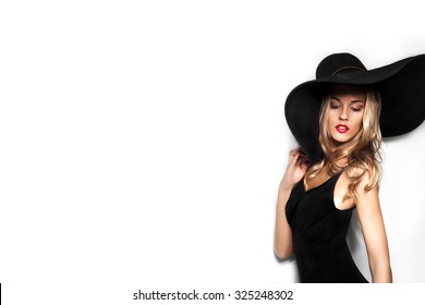 High Fashion Shot Of Blonde Woman With Curly Hair In Black Hat And Stylish Elegant Evening Dress Posing On White Studio Background