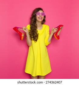 High Heels Is My Middle Name. Laughing Young Woman In Yellow Mini Dress And Pink Sunglasses Holding A Red High Heels. Three Quarter Length Studio On Pink Background.