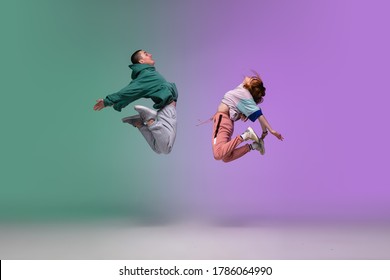 High jump. Boy and girl dancing hip-hop in stylish clothes on colorful gradient background at dance hall in neon. Youth culture, movement, style and fashion, action. Fashionable portrait. Street dance