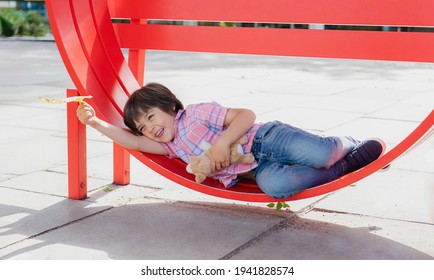 High Key Light Portrait Of Happy Little Boy Holding Autumn Leaf Lying On Red Chair, Adorable Child Playing With Outdoor Metal Chair, Kid Playing Outside, Positive Children Concept