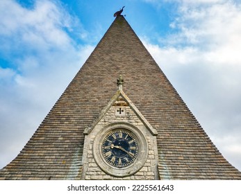 High level view of a large clock seen in a English clock tower with the church spire and metal weathercock atop.