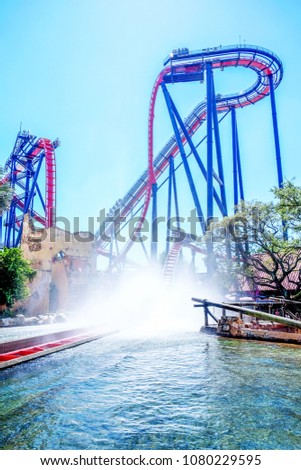 A High modern rollercoaster with a splash pool in the foreground the roller coaster ride is about to go into the splash pool blue sky and blue pool, metal construction roller coaster