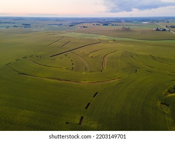 High Overhead View Of Lush Farm Field In Autumn.  Strategic Areas Unplanted Making Patterns In The Field. Hay Sky. Farm Buildings In The Distance With Trees. Small Rolling Hills Present.