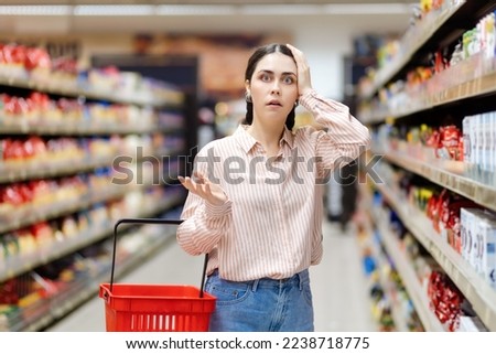 High price and inflation. Portrait of young shocked caucasian woman holds cart and takes hand to head. Showcase at background. Person forget to buy something. Concept of shopping in supermarket.