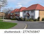 High quality housing development featuring beautiful detached houses with mixed roof tiles and varied facade materials, set within landscaped surroundings along a winding residential road. The scene shows a contemporary UK suburban estate with carefully planned architecture, varied housing designs, and integrated greenery, reflecting modern residential development and suburban planning.