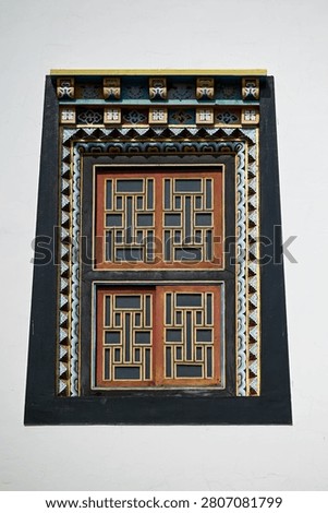 High resolution vertical closeup of a single trapezoid Tibetan window with carved geometric
lattice shutters and painted decorative frame on a white monastery wall