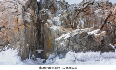 A high rock with many cracks, close-up. Flat stones are covered with snow. On the base there are ice splashes, icicles. In a narrow crevice, a man stands, with spread hands touched its walls. Baikal.