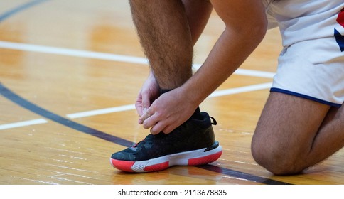 A High School Basketball Player Is On His Knee Tying His Shoes During A Game Indoors.