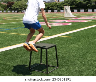 High school boy in the air while jumping on to a black medal plyo box on a green turf field at practice. - Powered by Shutterstock - Get 15% off with code: PIKWIZARD15