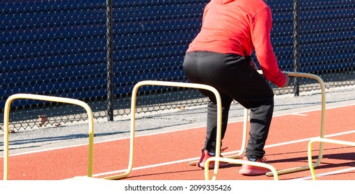 High School Boy Jumping Over Yellow Plastic Hurdles During Track Practice Landing On A Red Track Ready To Jump Again.