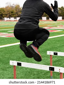 A High School Boy Is Jumping Over Track Hurdles On A Turf Field Wearing Sweatpants And Gloves On A Cold Day During Track Practice.