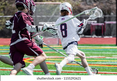 A high school boy lacrosse player is running down the field with the ball in his stick, being chased by the competition.