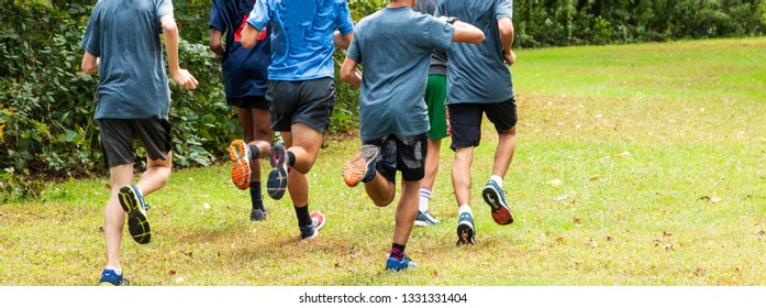 A High School Boys Cross Country Team Is Running Together On A Grass Path In A Local Park During Practice.