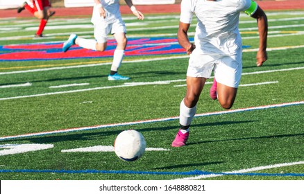 High School Boys Soccer Game With An Athlete Chasing Th Eball Down The Field Wearing A White Uniform.