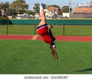 A High School Cheerleader Is Flipping And Twisting Updide Down In The Air While Warming Up For Pep Rally On A Gree Turf Field.