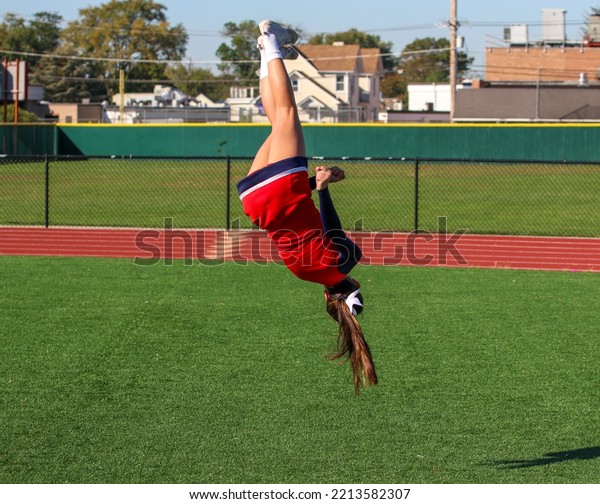 A high school cheerleader is flipping and twisting\
updide down in the air while warming up for pep rally on a gree\
turf field.