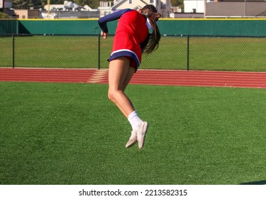 A High School Cheerleader Is Twisting Midair While Warming Up For Pep Rally On A Turf Field.