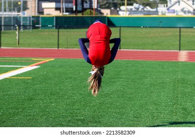 A High School Cheerleader Is Upside Down Flipping In The Air While Warming Up For Pep Rally On A Green Turf Field.