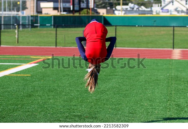 A high\
school cheerleader is upside down flipping in the air while warming\
up for pep rally on a green turf\
field.