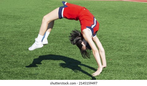 High school cheerleader warming up doing back tumbles and flips with her shadow on a green turf field before her performance.
